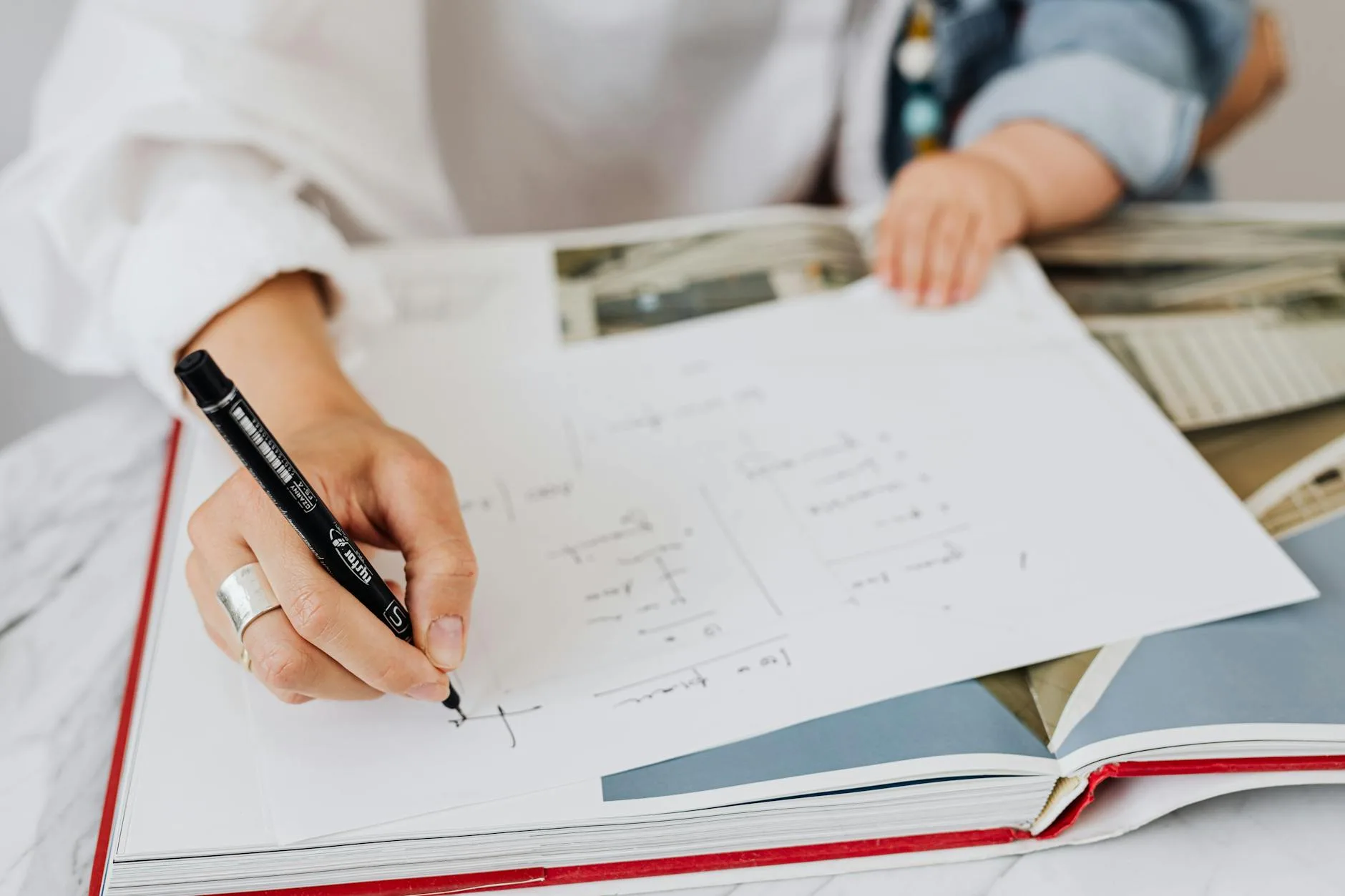 Dłoń pisząca na kartce przy książce, obok siedzi dziecko